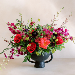Lush bouquet of red roses, pink peonies, and purple orchids in a dark pedestal vase on a pale surface.