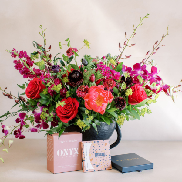 Vibrant flower arrangement in a dark pot with red, pink, and purple blooms, set on a light table with pink coffee and chocolate boxes nearby.