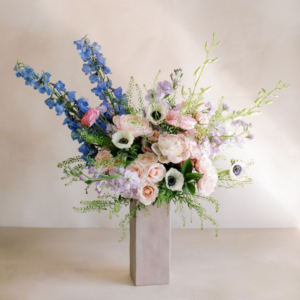 Pastel floral bouquet in a tall rectangular pale vase on a neutral surface, with pink roses and blue delphiniums.
