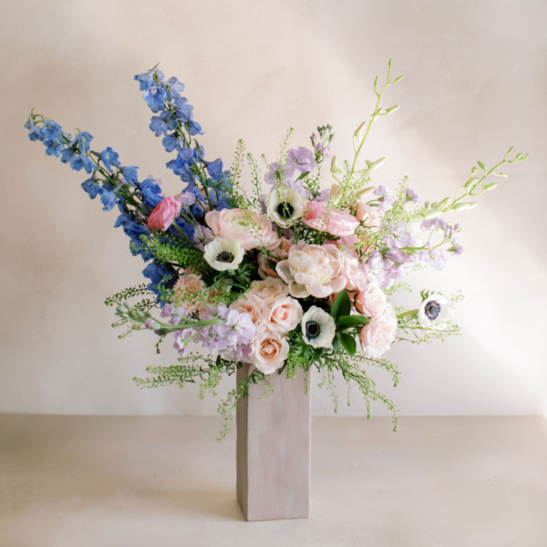 Pastel floral bouquet in a tall rectangular pale vase on a neutral surface, with pink roses and blue delphiniums.